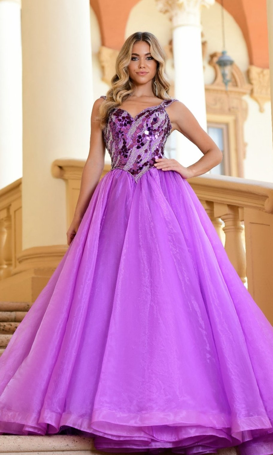 A woman stands on stairs wearing the Long Formal Dress 28579 by Ava Presley, a sleeveless purple A-line gown with a sequined, embroidered bodice and full tulle skirt. Elegant pillars and architectural details are in the background.